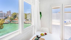 A room with a large window overlooking a body of water and buildings, a white table with a plate of fruit, and a chair.