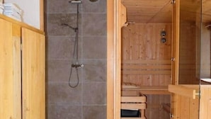 A wooden bathroom with a shower, a wooden bench, and a glass door leading to another room.