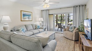A living room with a sofa, coffee table, and TV. There is a ceiling fan, a painting, and a view of the outdoors.