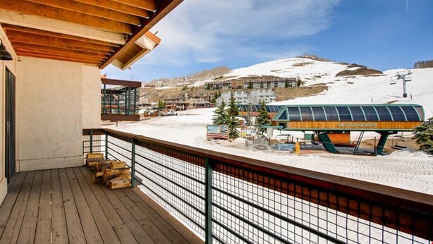 Una terraza de madera con baranda de metal, con vista a un paisaje nevado que incluye un telesilla y edificios al fondo.