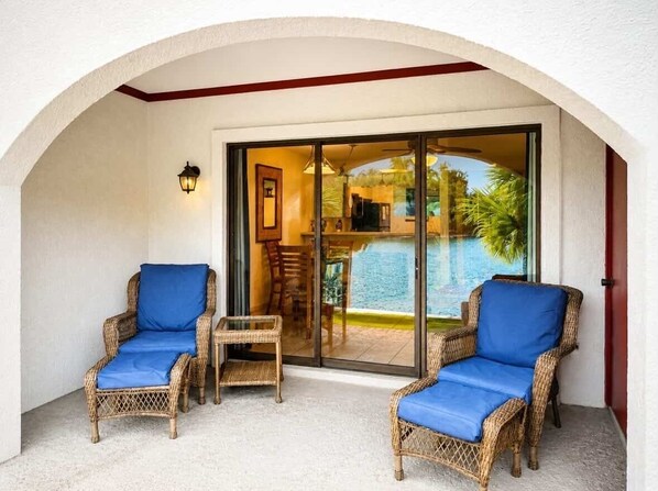 Un espace extérieur abrité avec des chaises en osier et une table, une porte vitrée donnant sur une piscine, et une bordure rouge le long du plafond.