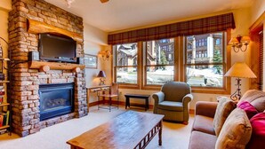 A cozy living room with a stone fireplace, comfortable seating, and large windows offering a view of the outside. - Deluxe, Classic Mountain Condo | Near Chairlifts (Crested Butte)
