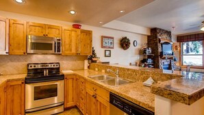 A kitchen with wooden cabinets, a stainless steel stove, granite countertops, a dishwasher, and a stone fireplace. - Deluxe, Classic Mountain Condo | Near Chairlifts (Crested Butte)