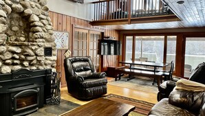 A cozy living room with a stone wall, leather furniture, a fireplace, and a balcony with a view. - 220’ WATERFRONT BEACH HOME ROCK RIVER WI PETS PIER POOLS FP FISHNG BOAT KAYAKS (Edgerton)