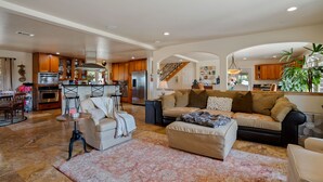 A spacious living room with a large beige sofa, a coffee table, and a patterned rug. There is a kitchen area with wooden cabinets and a refrigerator in the background.