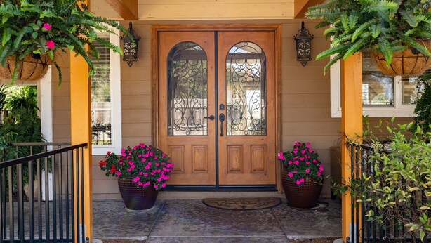 A wooden double door with decorative glass panels, flanked by large potted plants with pink flowers, and a black metal railing.