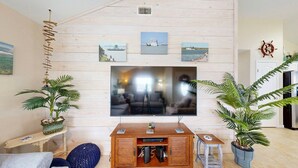 En stue med et fladskærms-tv monteret på en trævæg, et træbord, to potteplanter og et maleri af en sejlbåd over tv'et.