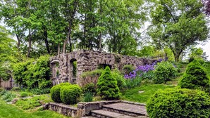 Un jardín con escalones de piedra que conducen a una edificación de piedra, rodeado de abundante follaje y plantas con flores.