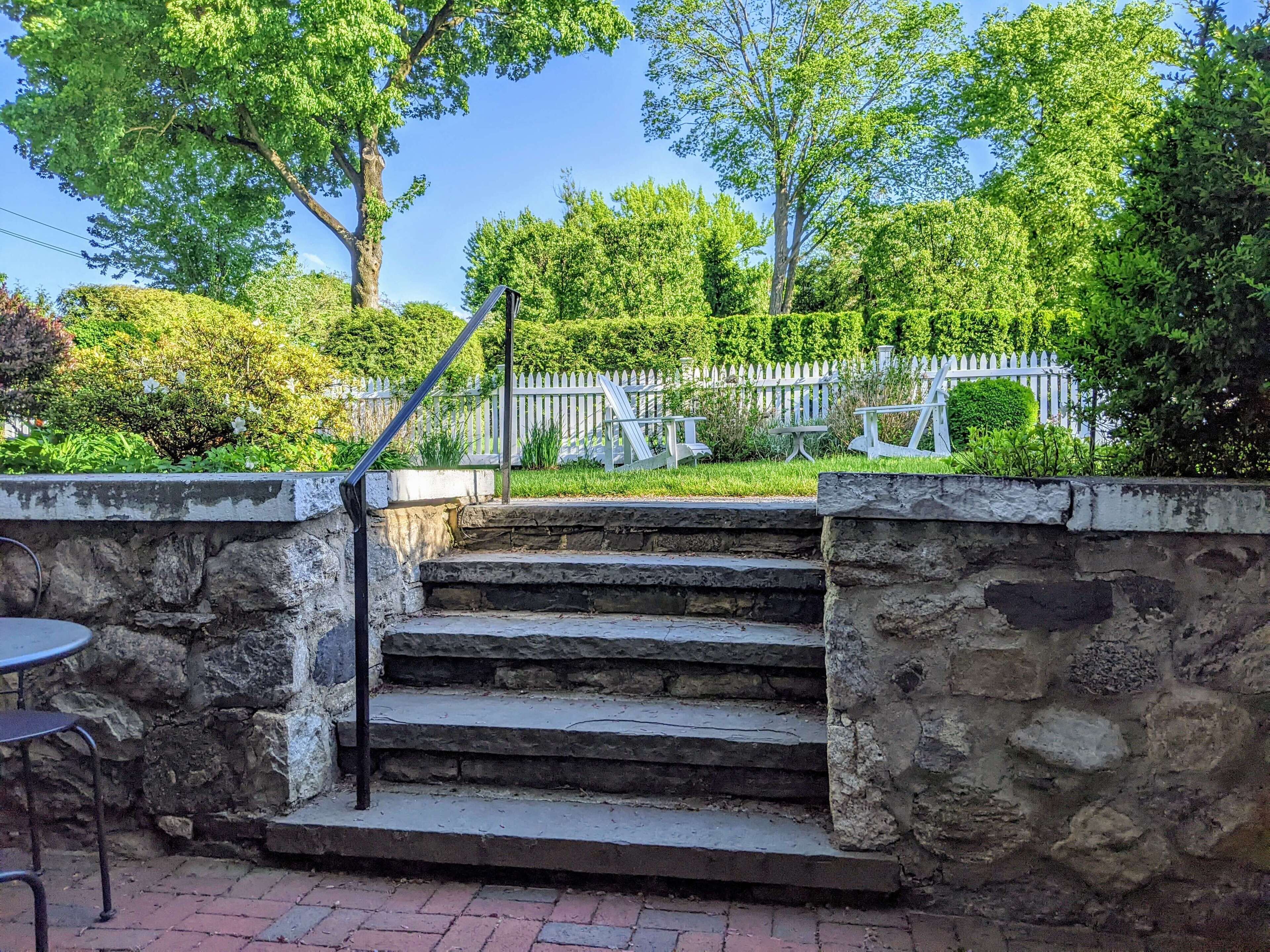 Un escalier en pierre mène à un jardin agrémenté d’une clôture blanche en bois et d’un espace aménagé pour s’asseoir à l’extérieur.