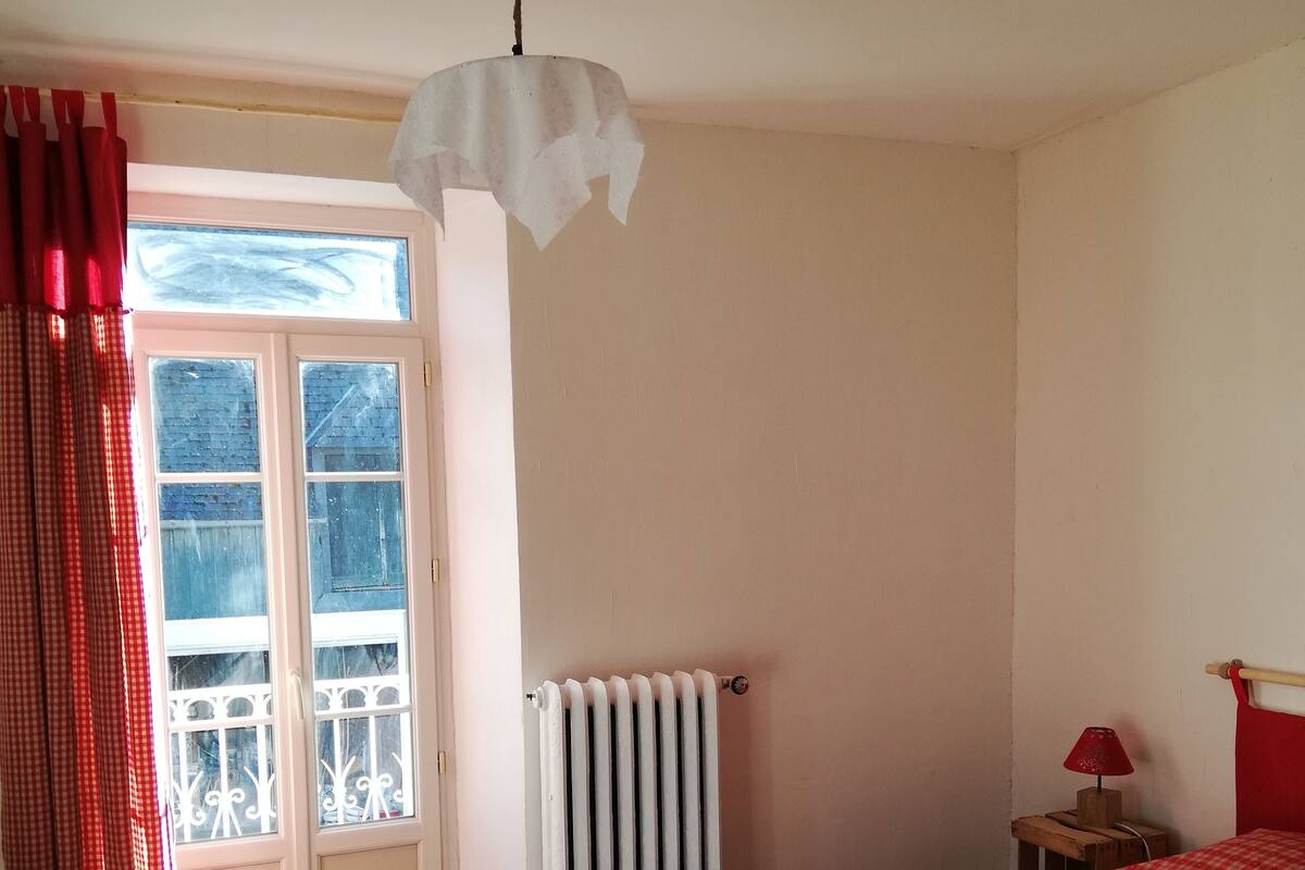 A bedroom with a wooden floor, a bed with a red checkered bedspread, a radiator, a small bedside table with a lamp, and a window with red curtains.