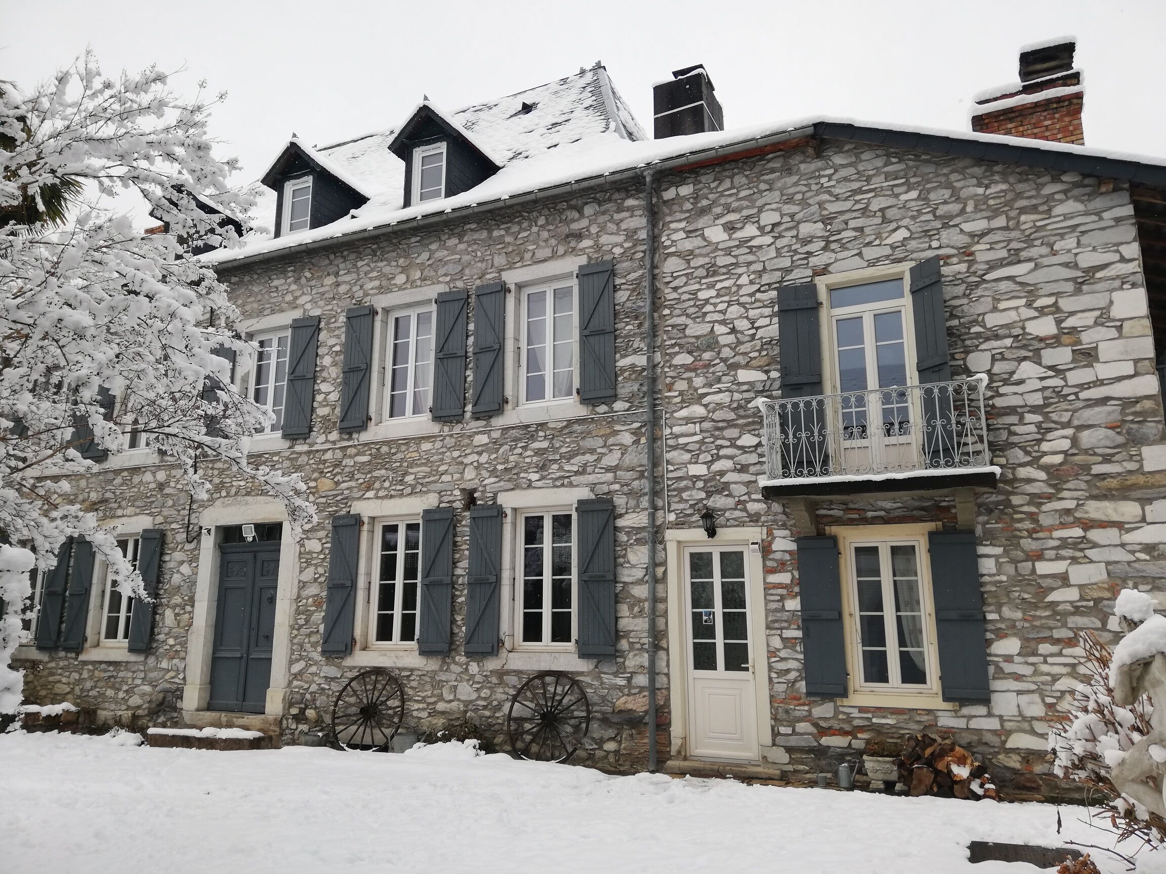 A stone building with multiple windows and shutters, a balcony with a metal railing, and snow on the roof and ground.