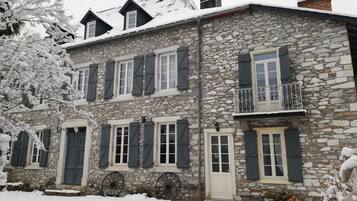 A stone building with multiple windows and shutters, a balcony with a metal railing, and snow on the roof and ground.