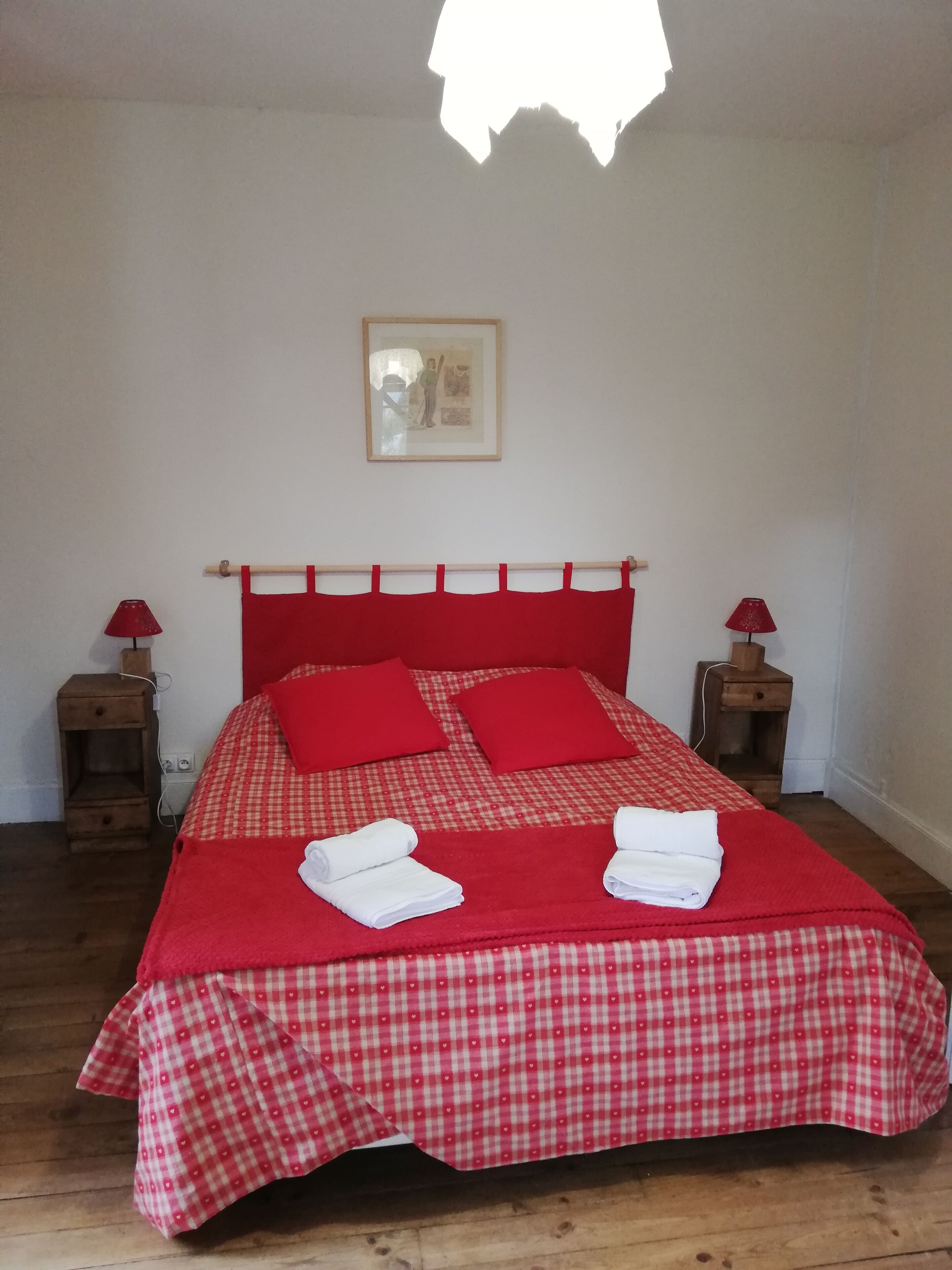 Una cama con una colcha y almohadas a cuadros rojos, dos mesitas de noche con lámparas, y un cuadro en la pared.