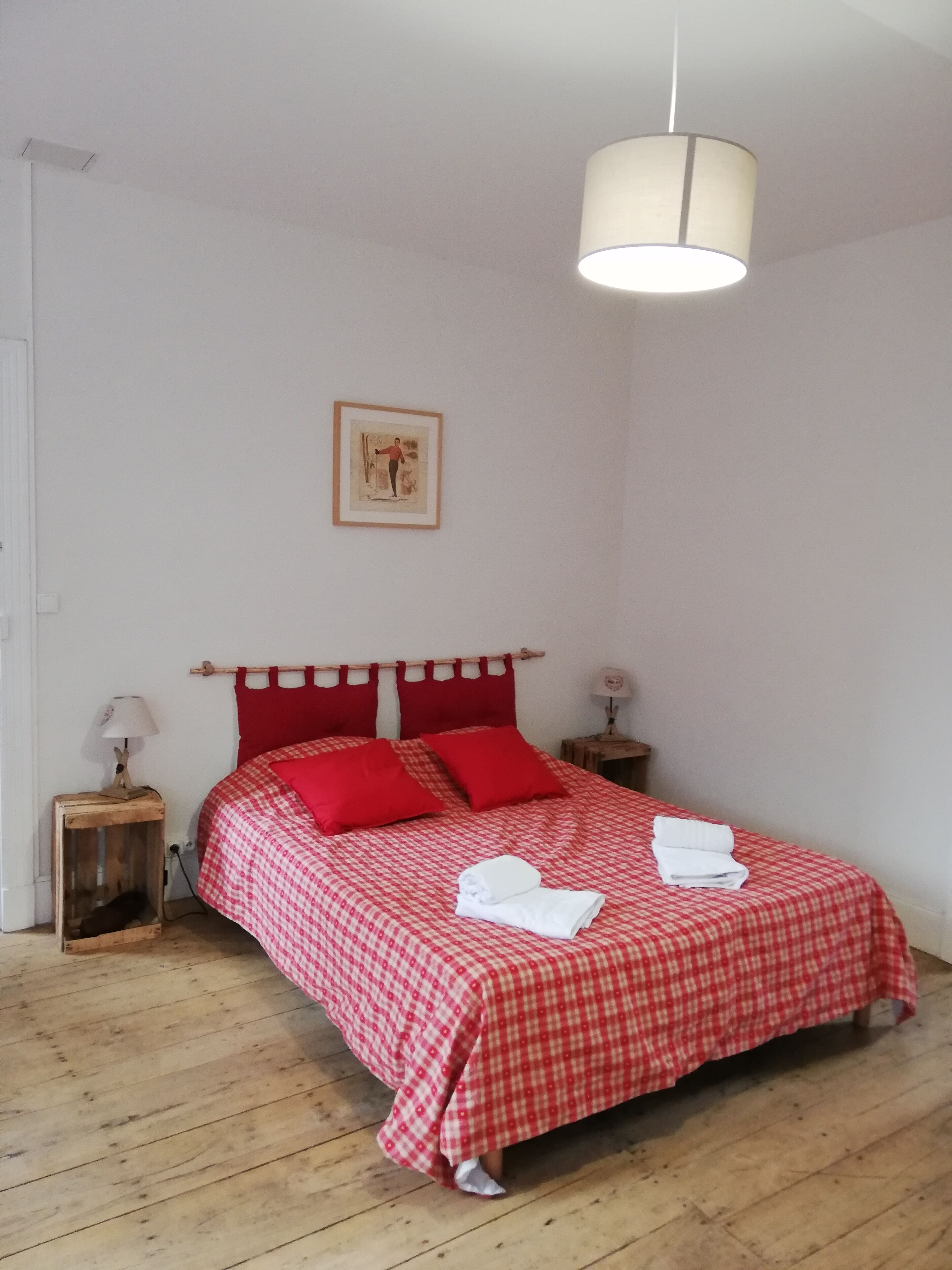 Un dormitorio con colcha a cuadros, almohadas rojas, cabecera de madera, dos mesitas de noche con lámparas y un cuadro enmarcado en la pared.