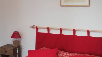 A bed with a red checkered bedspread and two red pillows, a wooden nightstand with a lamp, and a framed picture on the wall.