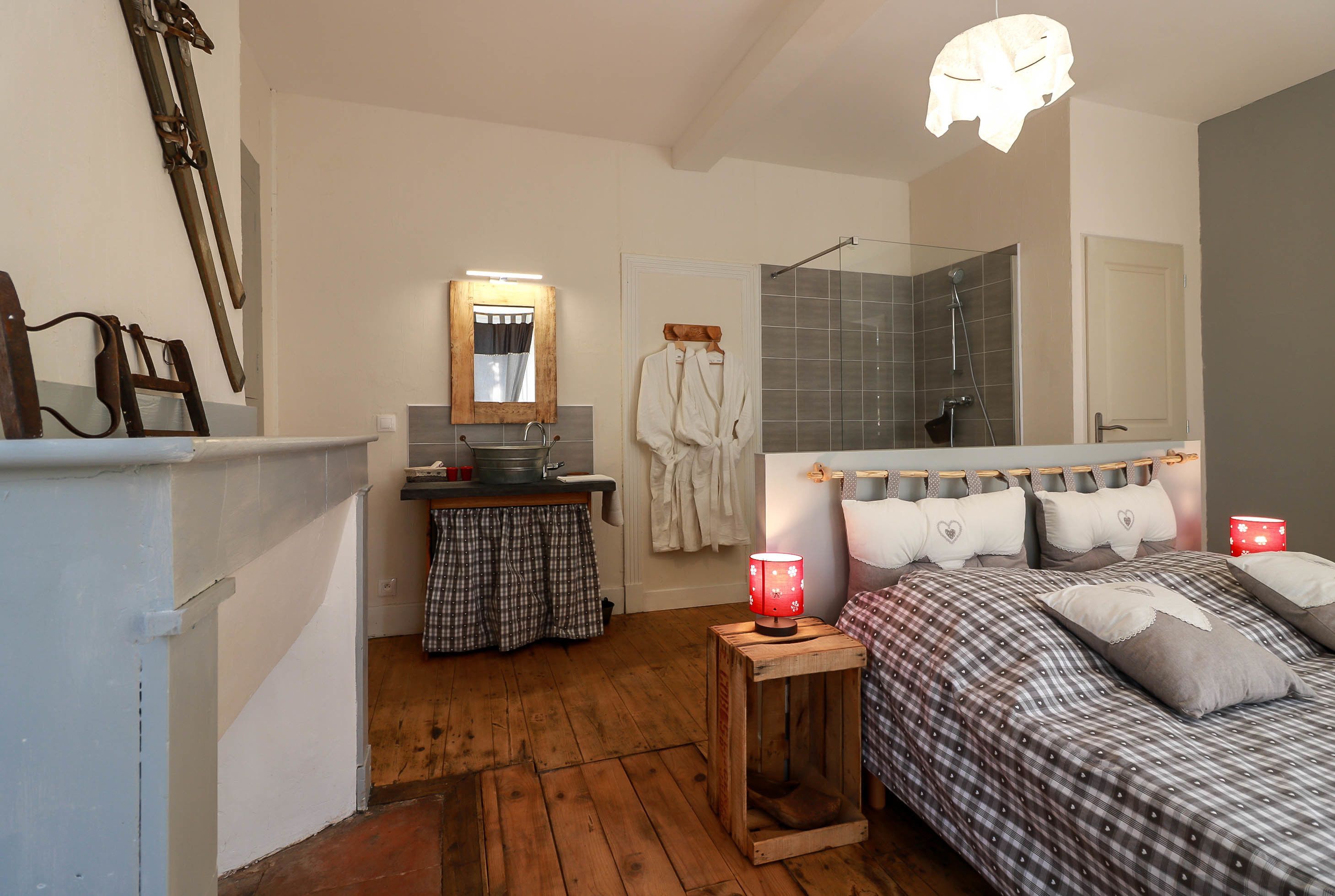 Un dormitorio con piso de madera, una cama con colcha a cuadros, una mesita de noche con lámpara, un espejo sobre la cama y una zona de ducha con puerta de cristal.