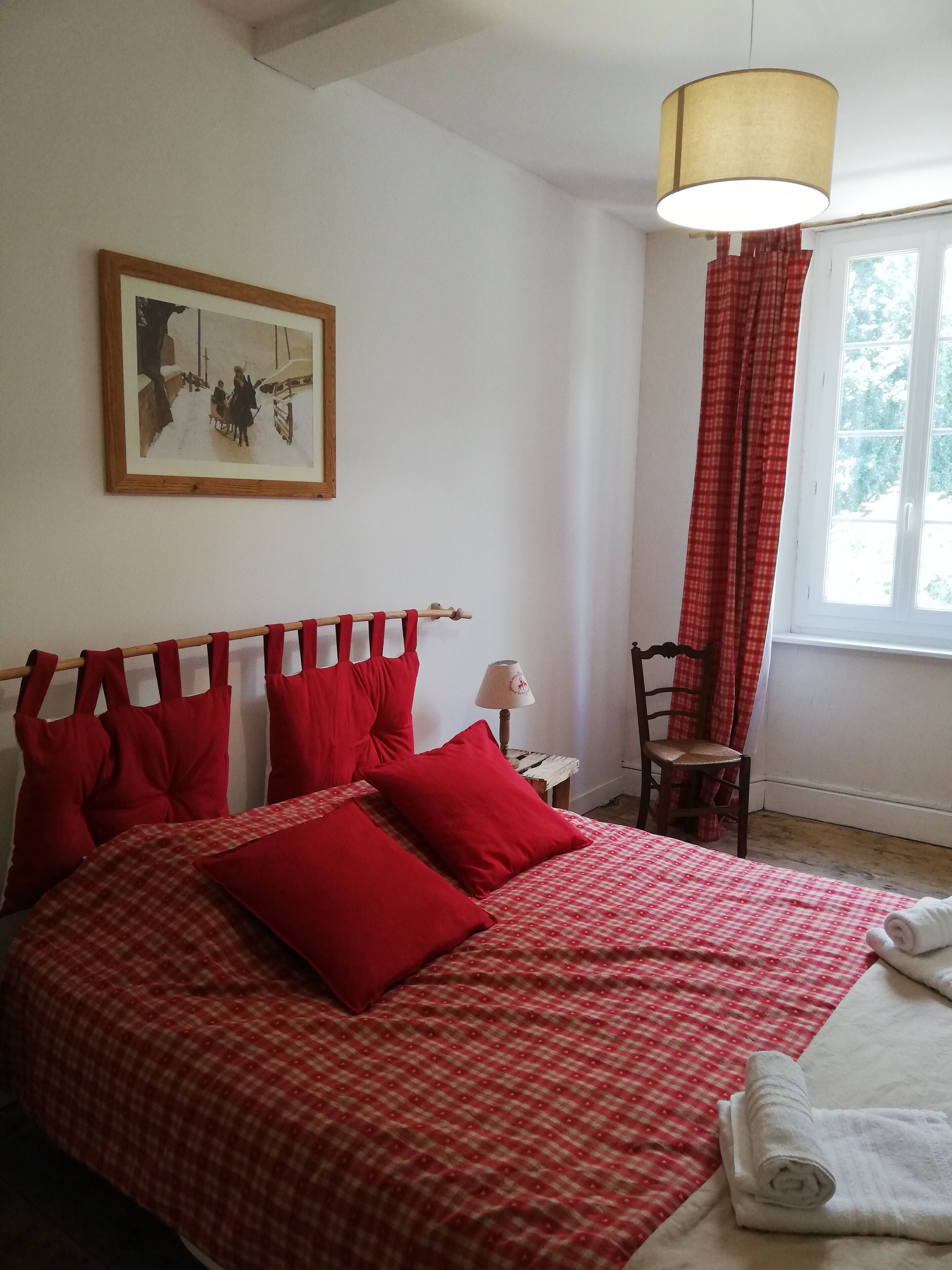 Une chambre à coucher avec un lit, des oreillers rouges, une tête de lit en bois, une petite table avec une lampe, une chaise, une fenêtre avec des rideaux rouges et un tableau encadré au mur.