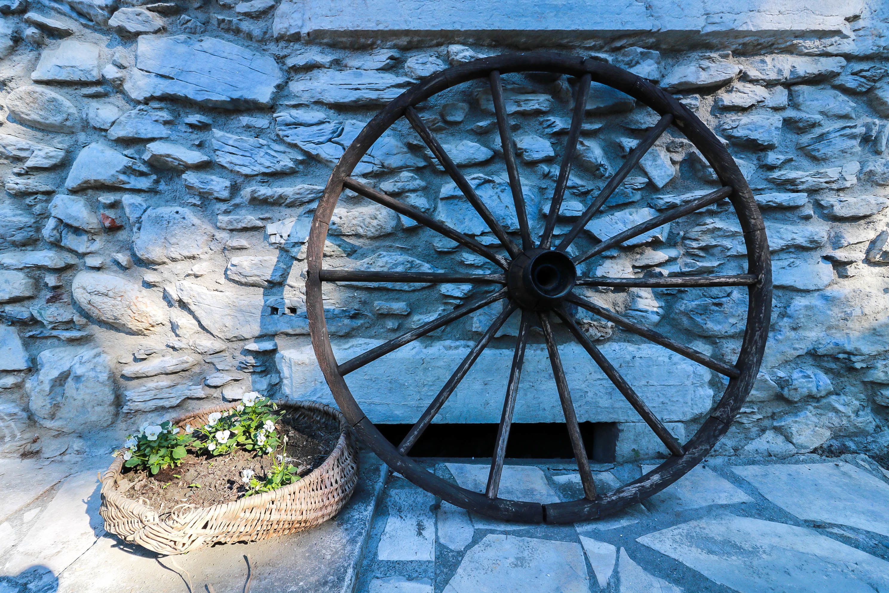Un muro de piedra con una rueda rústica grande y una cesta de mimbre con plantas.