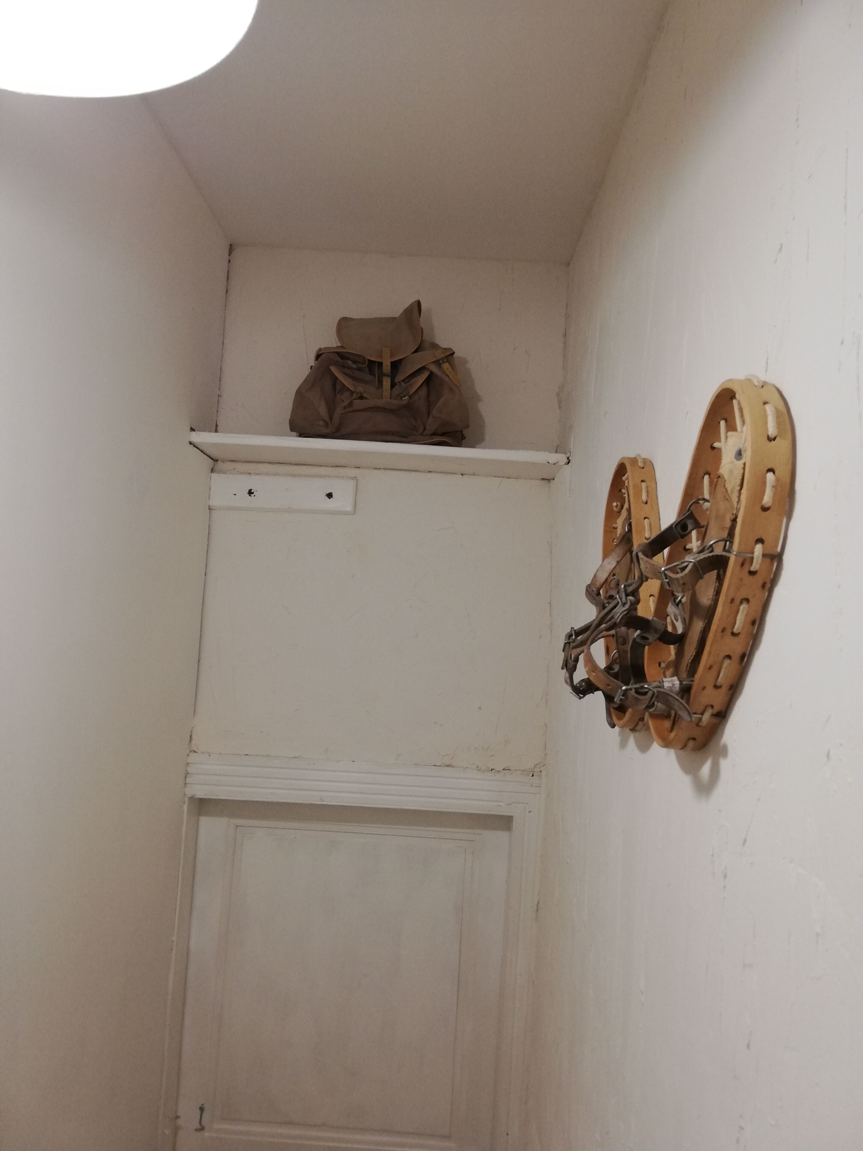 Una esquina de una habitación con una puerta blanca, una estantería con una bolsa marrón y un par de esquíes colgados en la pared.