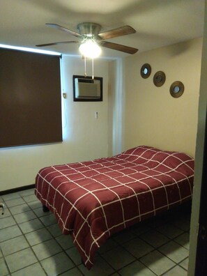 A single bed with a patterned bedspread in a room with a ceiling fan, a window with blinds, and a wall-mounted air conditioning unit.