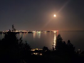 Una vista nocturna de una superficie acuática tranquila que refleja las luces de la ciudad, con una luna llena destacada en el cielo.