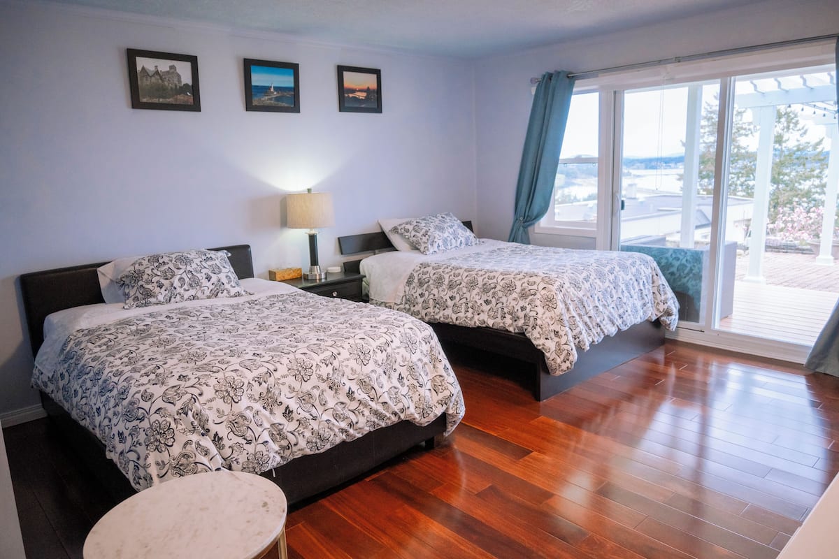 A bedroom with two beds, a nightstand, a lamp, and a view of the outdoors through a large window.
