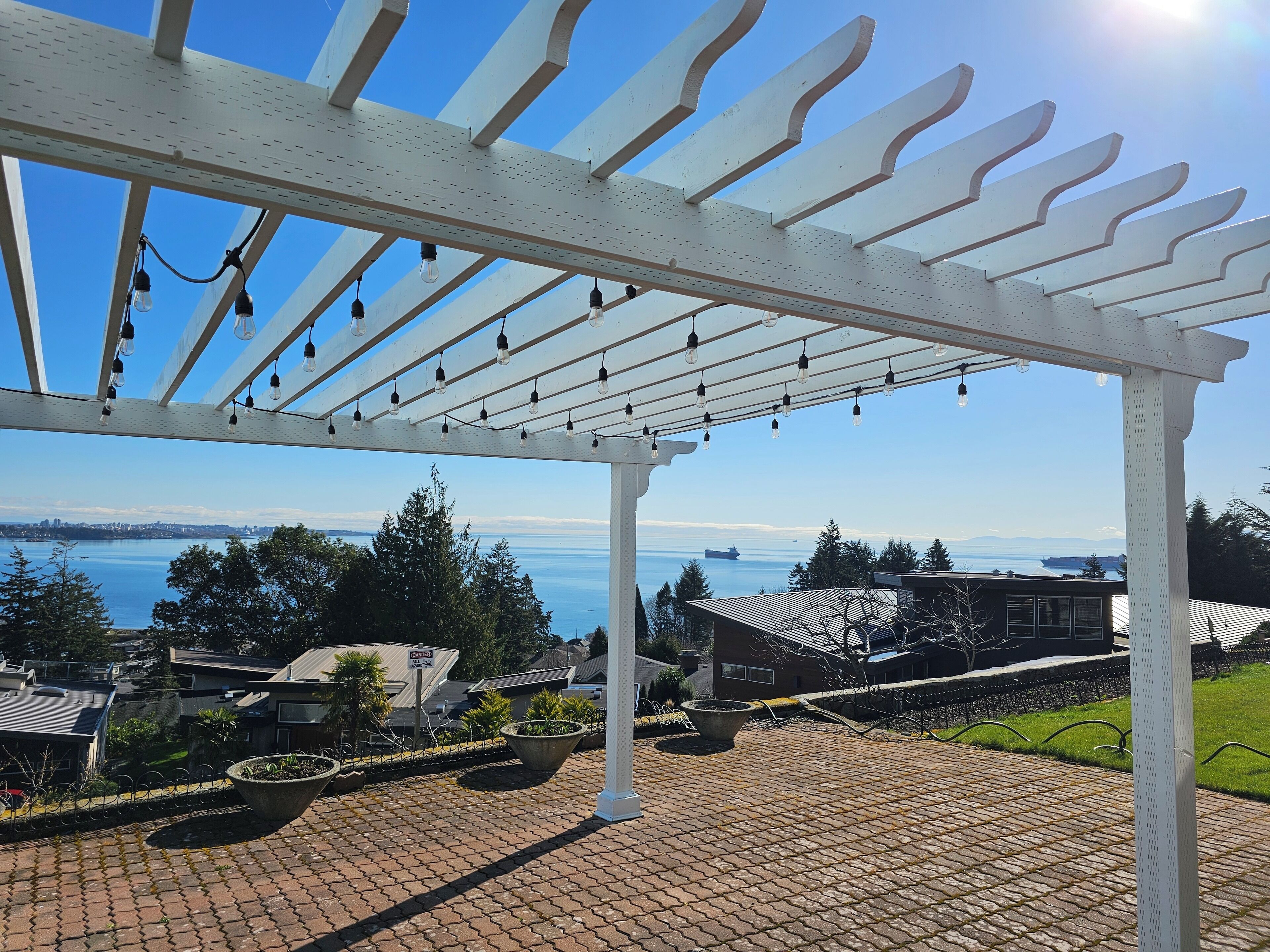 Un espacio al aire libre cubierto con una pérgola y luces colgantes, con vista a una masa de agua y edificios residenciales.