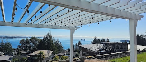 Un espacio al aire libre cubierto con una pérgola y luces colgantes, con vista a una masa de agua y edificios residenciales.