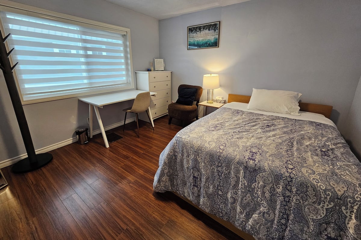 A hotel room with a wooden floor, a bed with a patterned bedspread, a desk with a chair, a dresser, and a lamp.