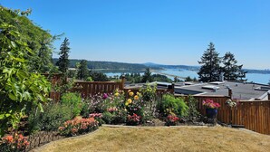 A backyard with a wooden fence, a variety of flowering plants, and a view of a lake and city skyline. - HOV B&B House -Hospitality Ocean view Victoria- (Victoria)