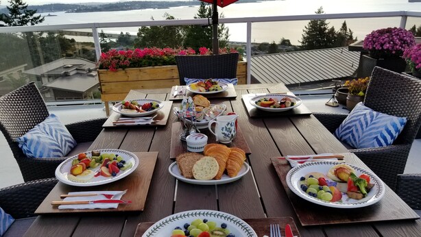 A wooden deck with outdoor dining tables set up with plates of food, cutlery, and wicker chairs. - HOV B&B House -Hospitality Ocean view Victoria- (Victoria)