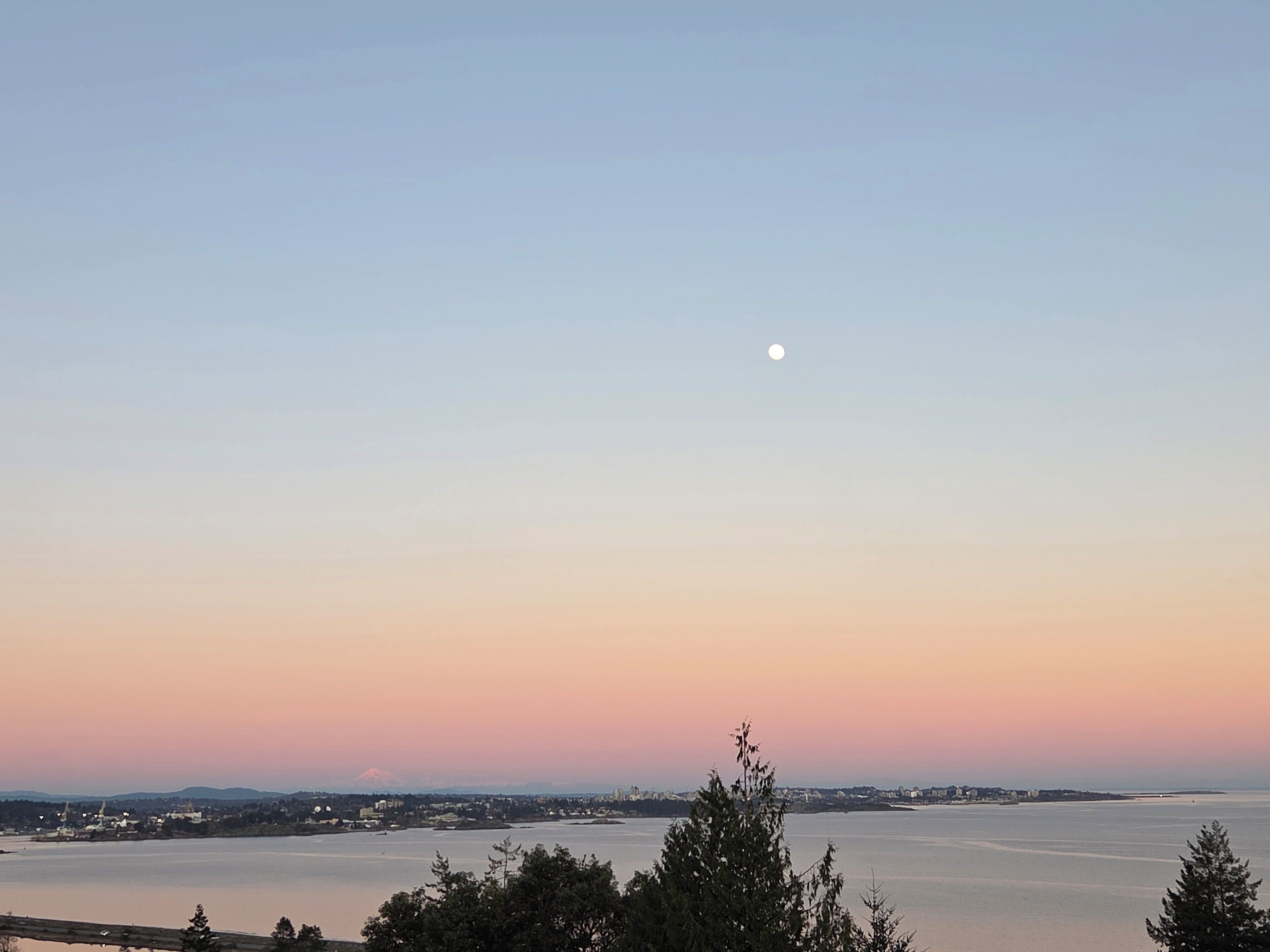 A coastal cityscape at dusk with a visible moon, calm water, and distant mountains.