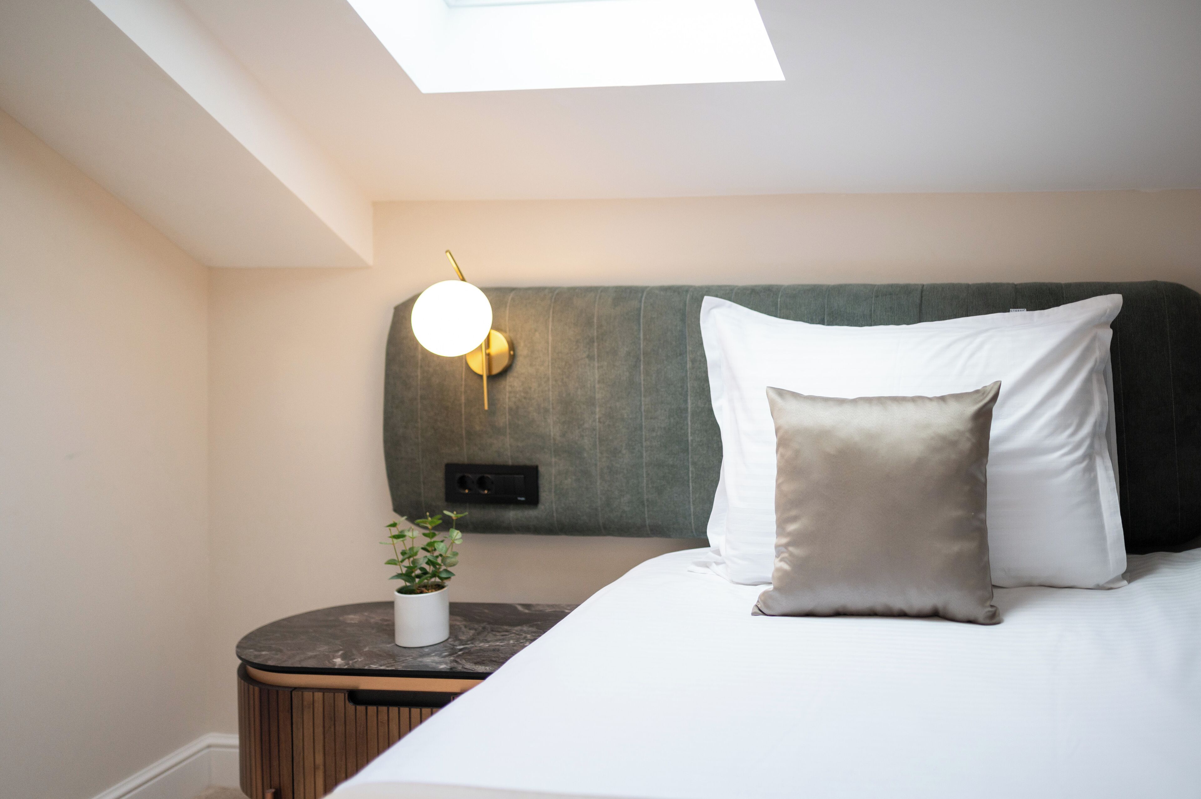 A hotel room with a skylight, a bed with white and beige pillows, a small round table with a plant, and a wall-mounted light fixture.