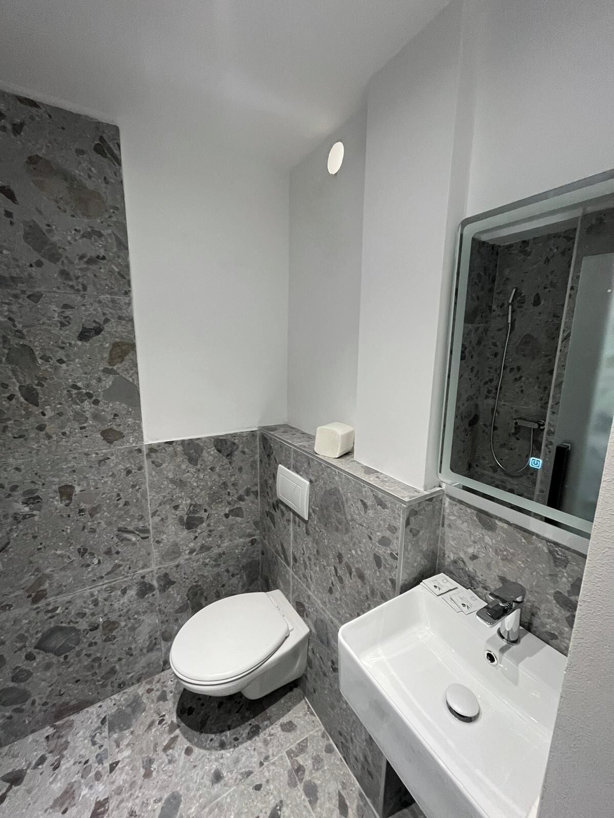 A small bathroom with a toilet, sink, and mirror. The walls are covered in gray stone tiles.