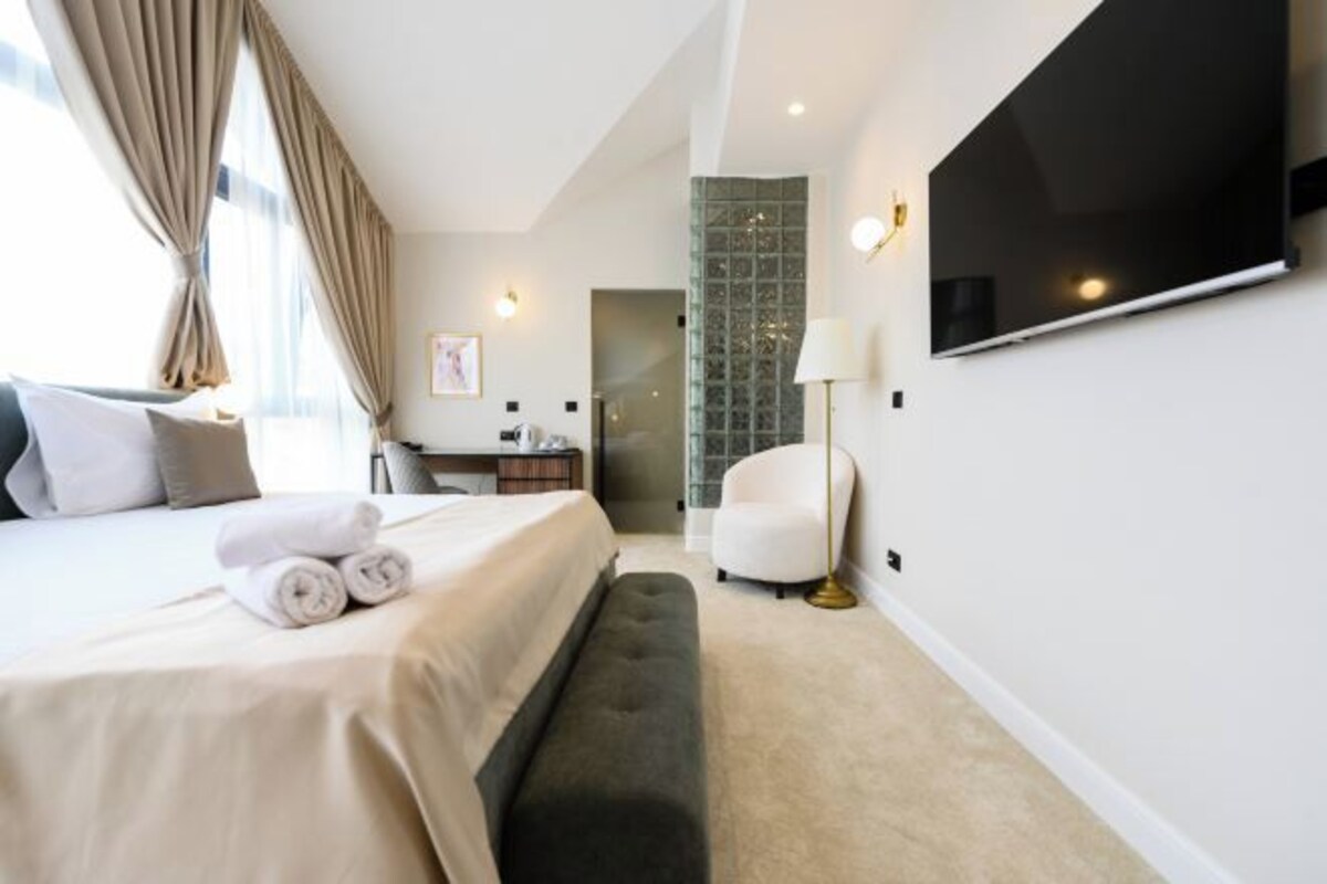 A modern hotel room with a large bed, a flat-screen TV mounted on the wall, a desk, a chair, and a bench.