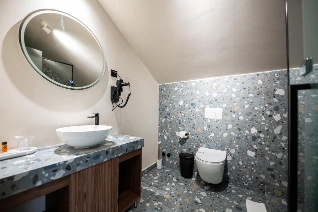 Een badkamer met een ronde spiegel, een witte wastafel, een toilet en een prullenbak.