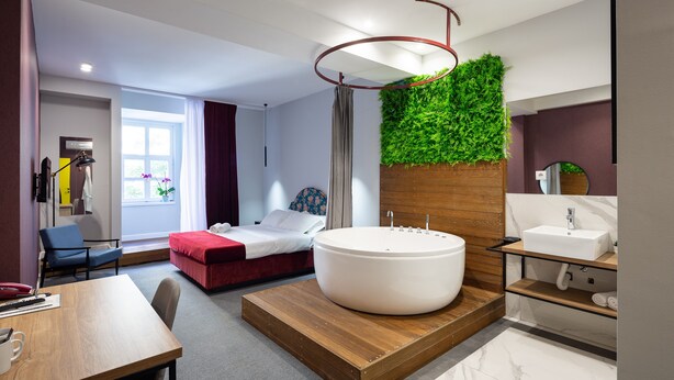 Una habitación de hotel moderna con una bañera grande, una plataforma de madera, una pared verde, un tocador de madera y una cama con ropa de cama roja.