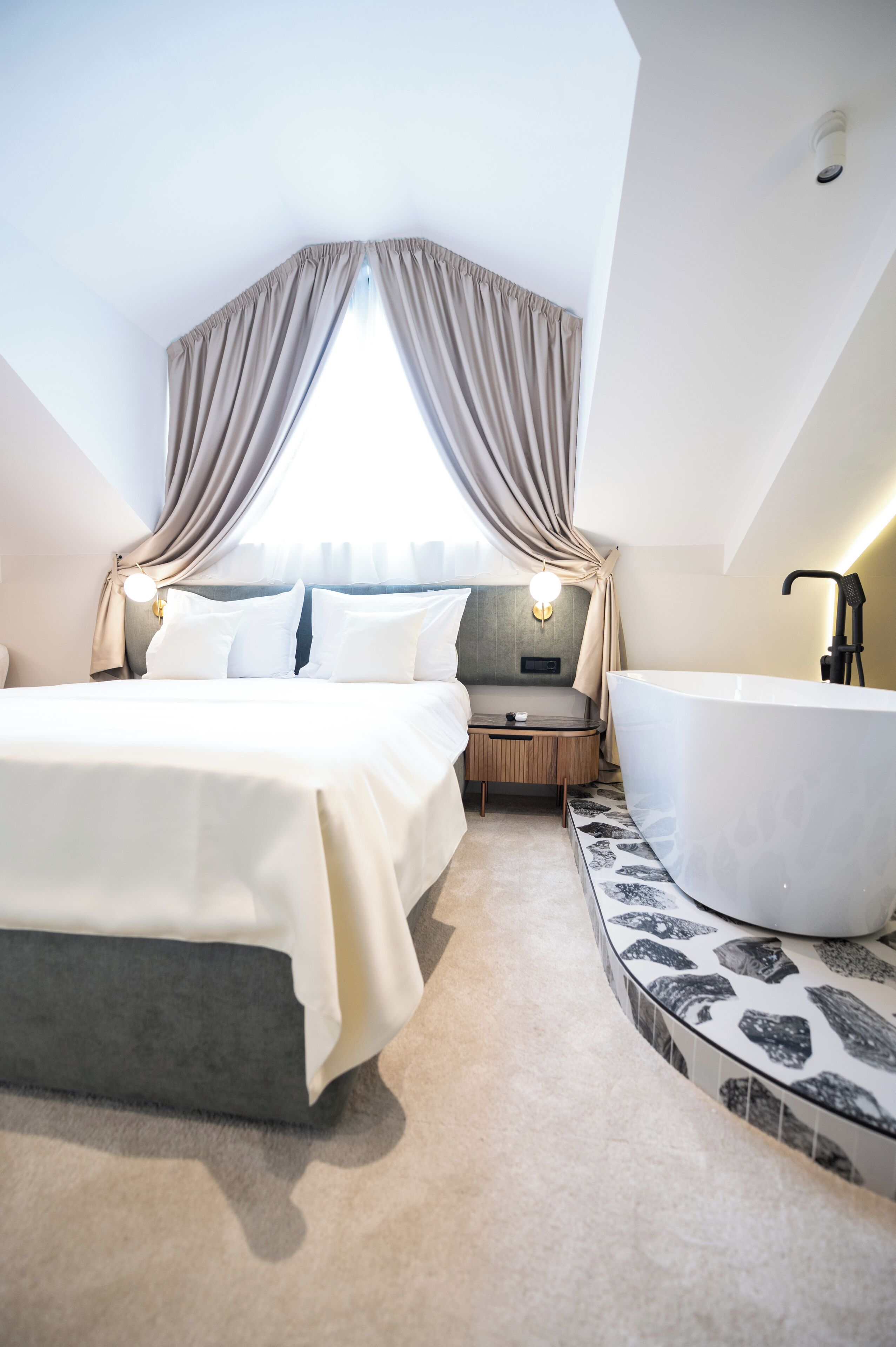 Una habitación de hotel moderna con una cama grande, una zona de estar separada, una bañera y vistas a la ciudad a través de una ventana con cortinas.