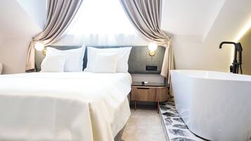 Una habitación de hotel moderna con una cama grande, una zona de estar separada, una bañera y vistas a la ciudad a través de una ventana con cortinas.