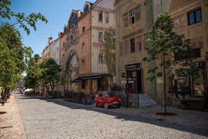 Une rue pavée bordée de bâtiments historiques, une voiture rouge d’époque garée devant un restaurant et un petit arbre au premier plan.