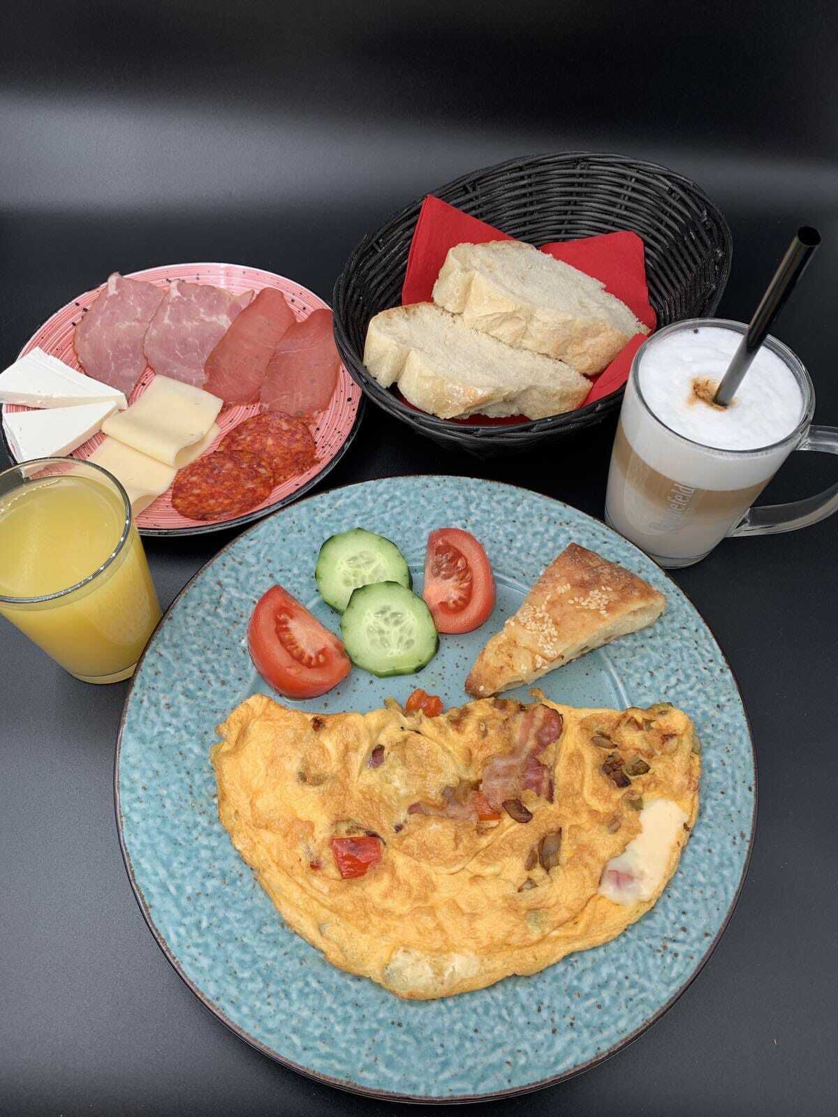 A breakfast plate with eggs, tomatoes, cucumbers, and bread, accompanied by a drink and a basket of meats and cheeses.