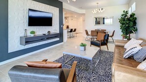 Una sala de estar moderna con un televisor de pantalla plana, una zona de comedor con mesa y sillas, y una planta grande.