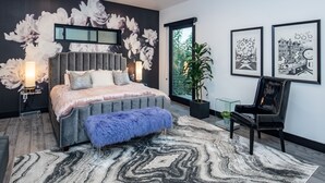 Una camera da letto moderna con un letto grande, un poggiapiedi blu soffice, una sedia nera e una grande finestra.