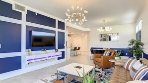 A modern living room with a flat-screen TV, comfortable seating, a coffee table, and a dining area in the background.