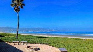 Ein gepflasterter Weg mit Bänken, einer Feuerstelle und einer Palme, mit Blick auf einen Strand und entfernte Berge.