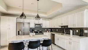 Una cocina moderna con armarios blancos, una isla central con taburetes de bar, electrodomésticos de acero inoxidable y salpicadero con mosaicos negros.