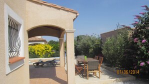 A patio with a table and chairs, a covered seating area, and a view of a garden with yellow flowers and a building in the background. - Stylish modern villa with private heated pool in scenic Trebes, near Carcassonne (Trèbes)