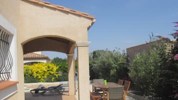 Une terrasse avec une table et des chaises, un espace salon couvert et une vue sur un jardin avec des fleurs jaunes et un bâtiment en arrière-plan.