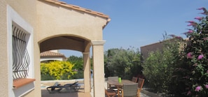 Une terrasse avec une table et des chaises, un espace salon couvert et une vue sur un jardin avec des fleurs jaunes et un bâtiment en arrière-plan.