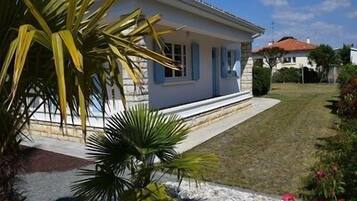 Une maison de plain-pied à l’extérieur blanc et aux bordures bleues, entourée d’une pelouse bien entretenue et d’une variété de plantes.